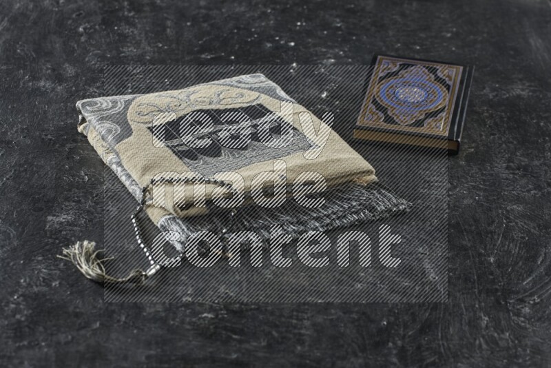 A folded prayer mat with different elements such as quran and prayer beads on black textured background