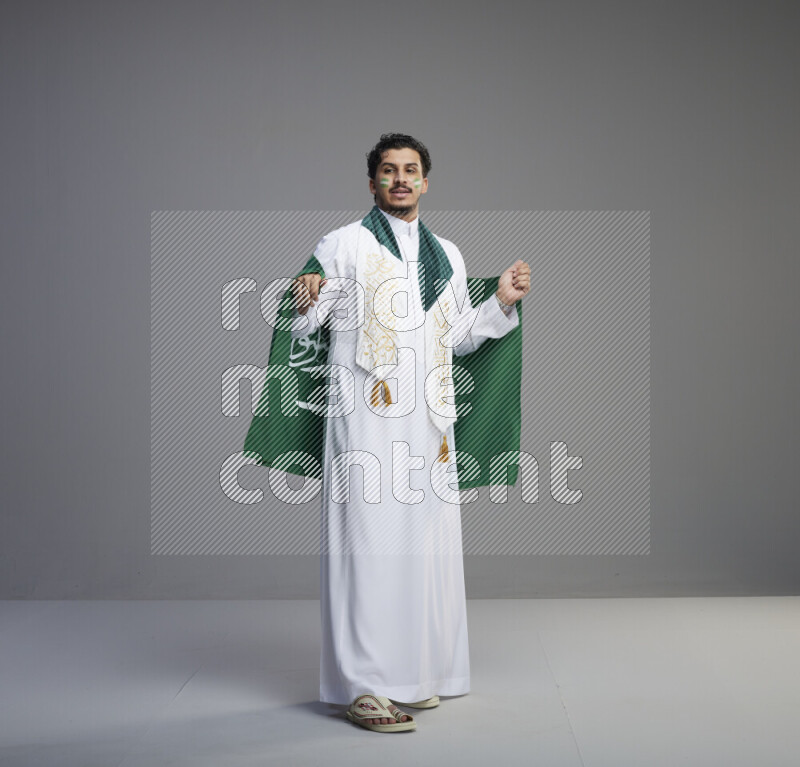 A Saudi man standing wearing thob with face painting wrapping Saudi flag scarf and holding big Saudi flag on gray background