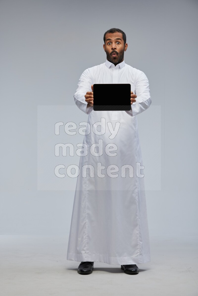 Saudi man wearing Thob standing showing tablet to camera on Gray background