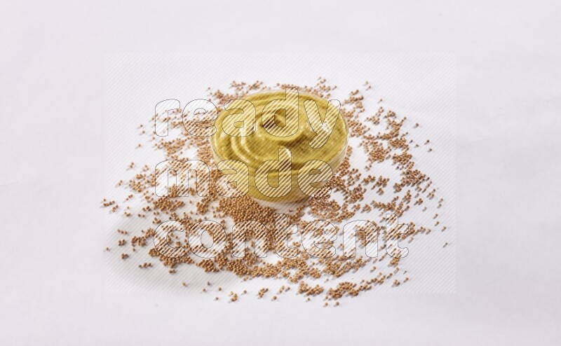 A glass bowl full of mustard paste with mustard seeds underneath on white flooring
