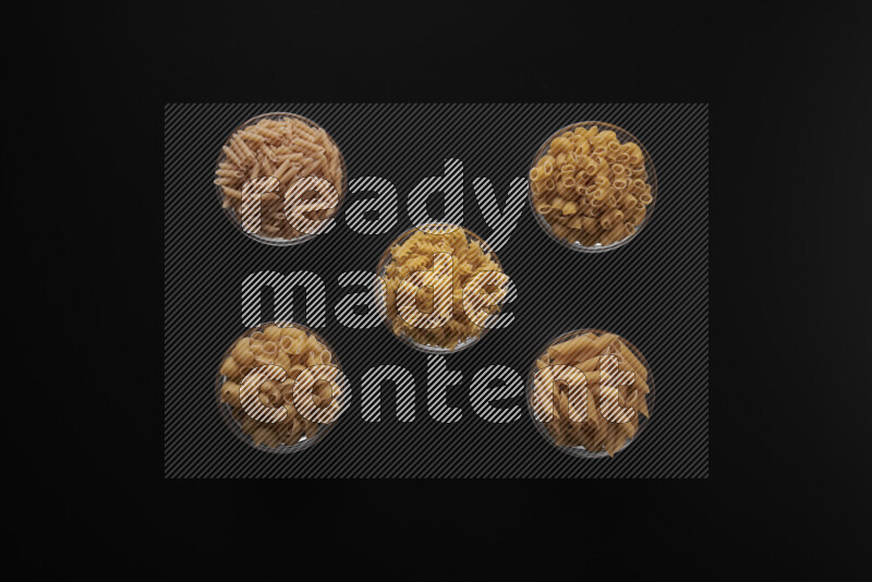 Different pasta types in 5 glass bowls on black background