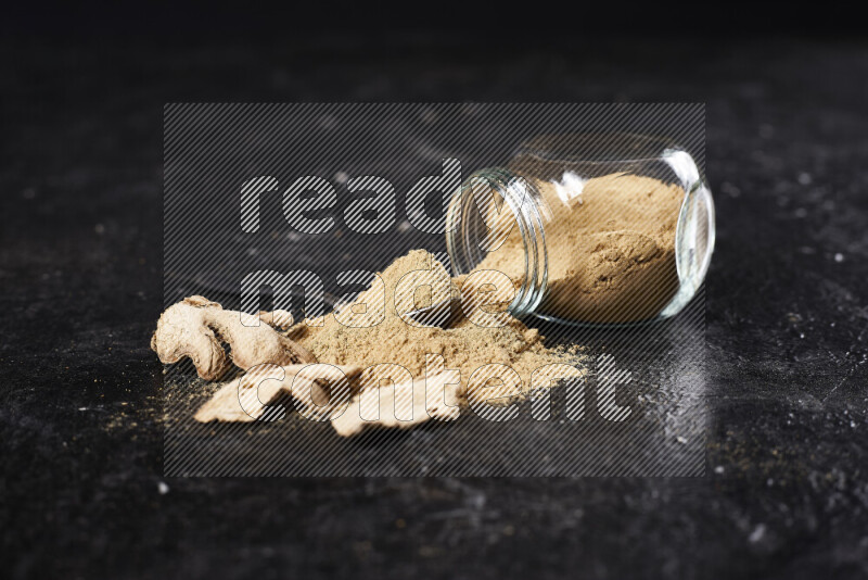 A glass jar full of ground ginger powder flipped with some spilling powder on black background