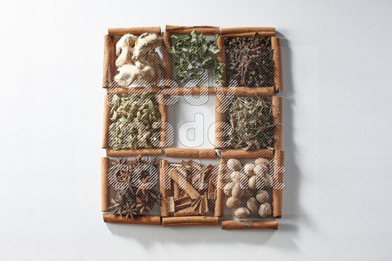 9 squares of cinnamon sticks, the middle square is empty and surrounded with dried mint, dried ginger, cardamom, star anise, cinnamon, nutmeg, dried basil and cloves on white flooring