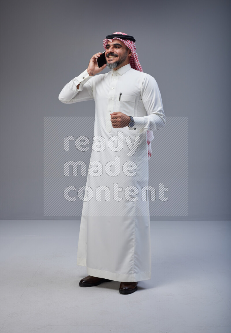 Saudi man Wearing Thob and red Shomag standing talking on phone on Gray background