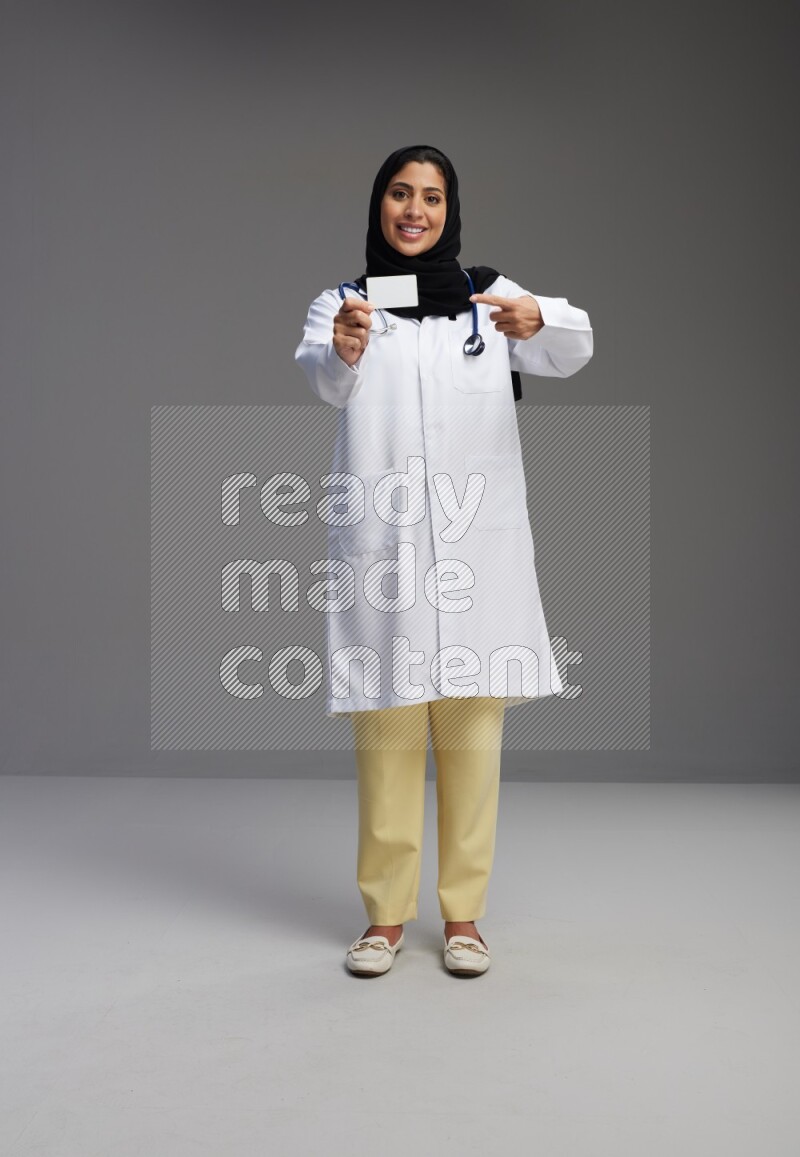 Saudi woman wearing lab coat with stethoscope standing holding ATM card on Gray background