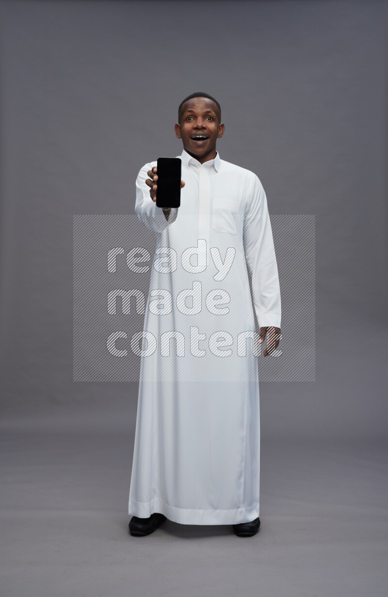 Saudi man wearing thob standing showing phone to camera on gray background