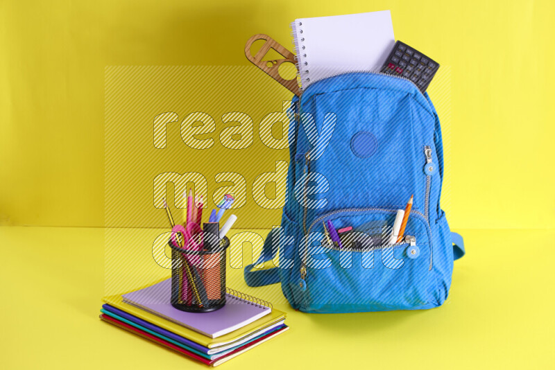 A school bag with assorted school supplies in and beside it on yellow background