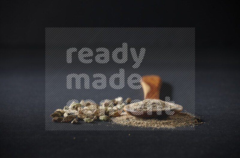 مغرفة خشبية ممتلئة ببودرة الهيل وحبوب الهيل بجانبها على خلفية سوداء