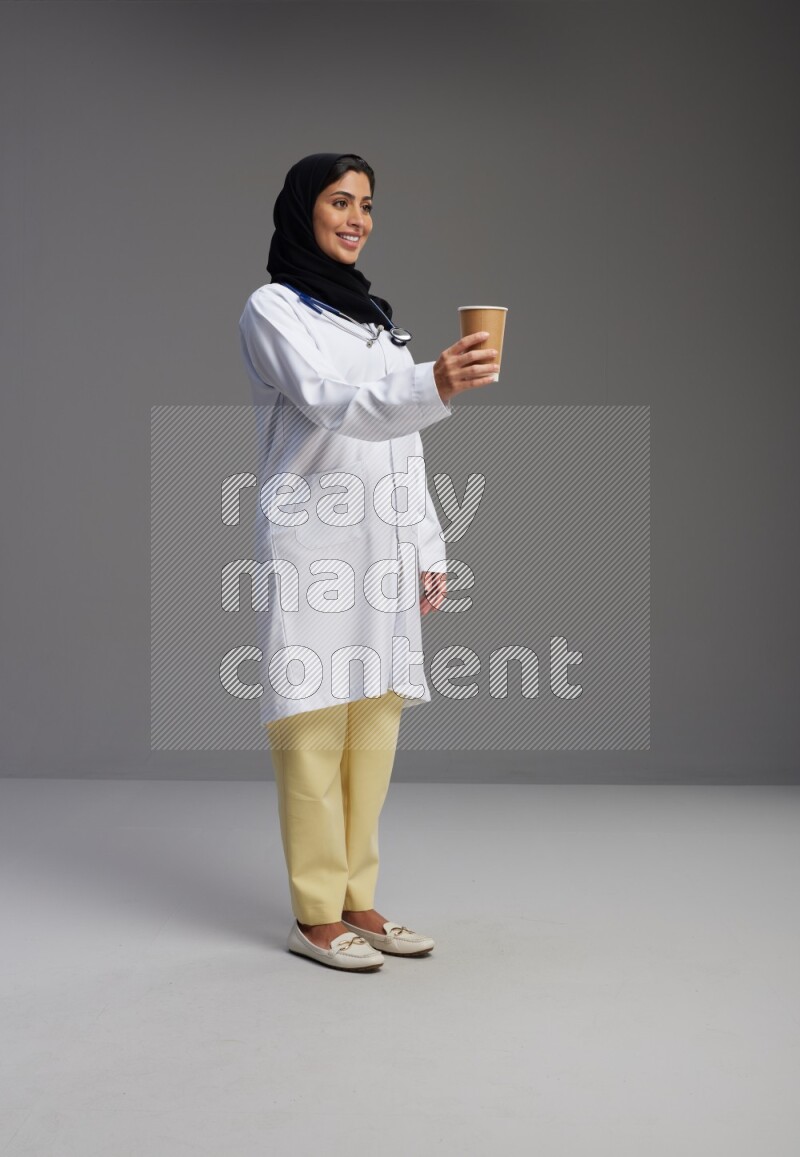 Saudi woman wearing lab coat with stethoscope standing holding paper cup on Gray background