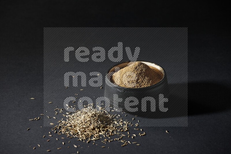 وعاء فخاري أسود ممتلئ ببودرة الكمون وحبوب الكمون متناثرة على خلفية سوداء