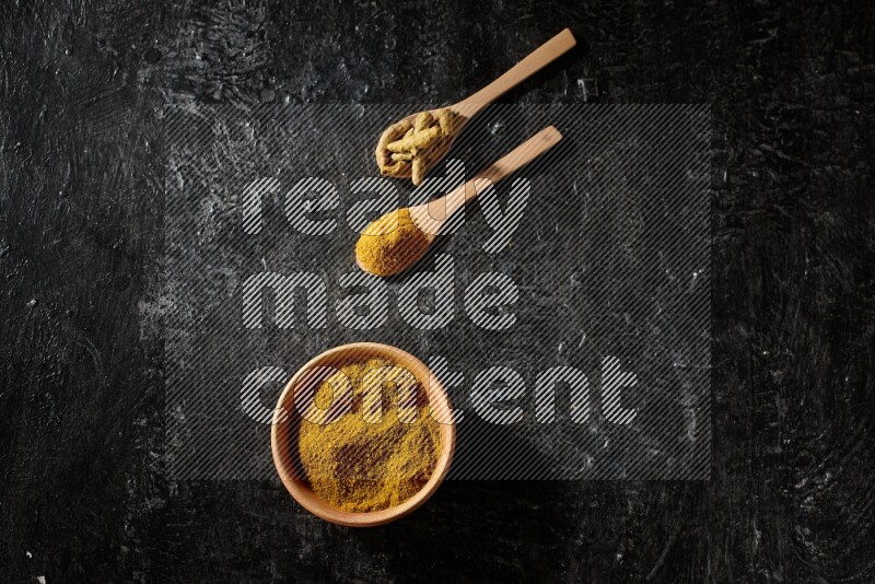 A wooden bowl full of turmeric powder and 2 wooden spoons full of dried turmeric finger and turmeric powder on textured black flooring