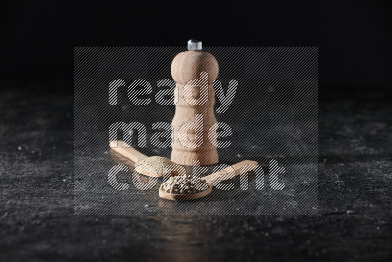 2 wooden spoons one full of white pepper powder and the other with pepper beads and a wooden pepper mill on textured black flooring