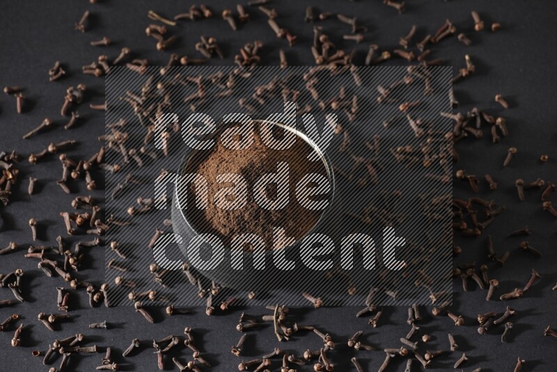 A black pottery bowl full of cloves powder and cloves whole grains on a black flooring