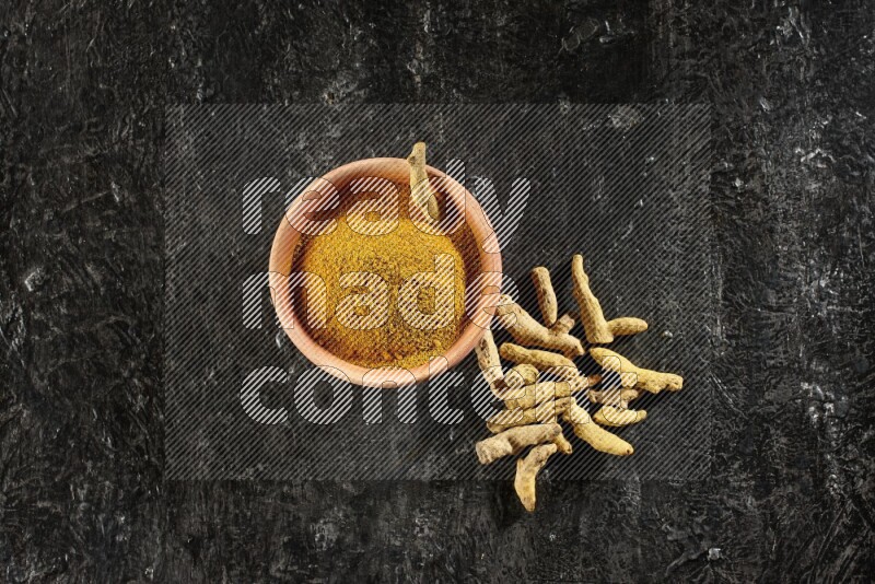 A wooden bowl full of turmeric powder with dried turmeric whole fingers on textured black flooring