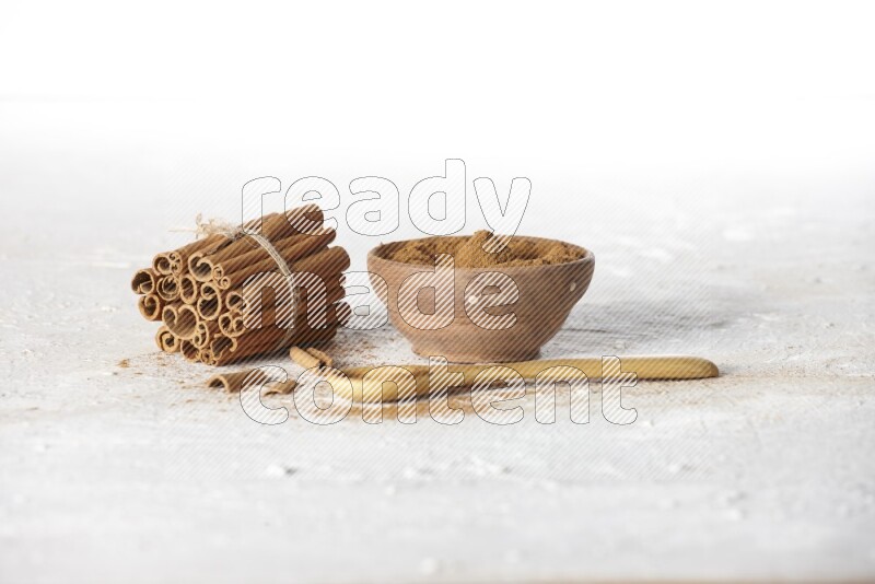 Cinnamon sticks stacked and bounded beside a wooden bowl full of cinnamon powder and a wooden spoon full of powder on white background