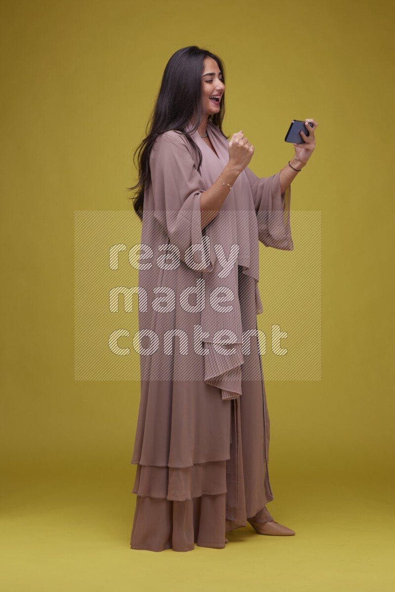 A woman Playing a Game on her smartphone on a Yellow Background wearing Brown Abaya