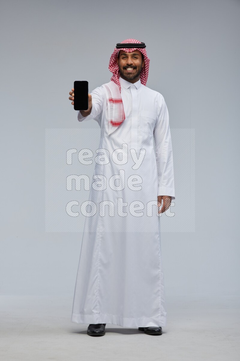Saudi man Wearing Thob and shomag standing showing phone to camera on Gray background