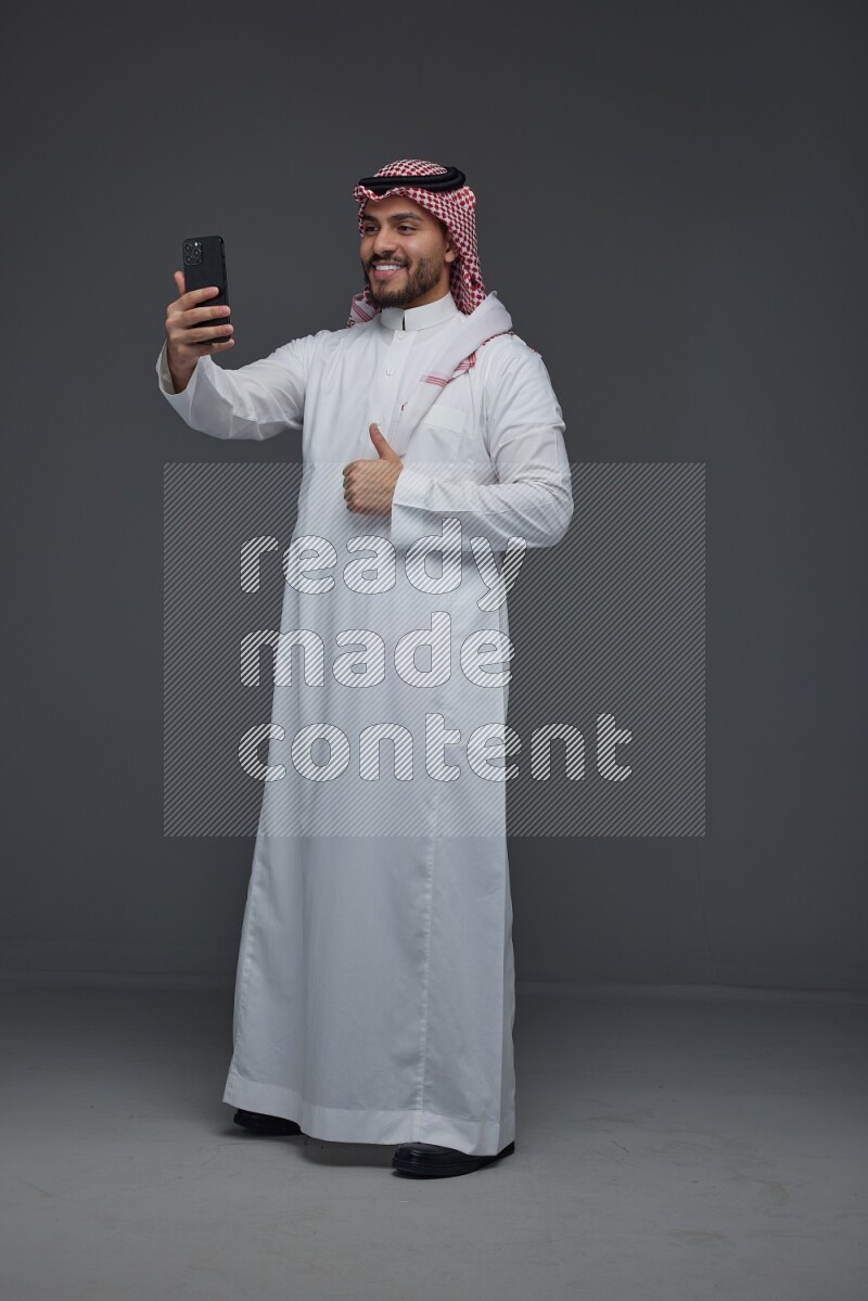 A Saudi man wearing Thobe and Shamgh taking selfie with his phone eye level on a gray background