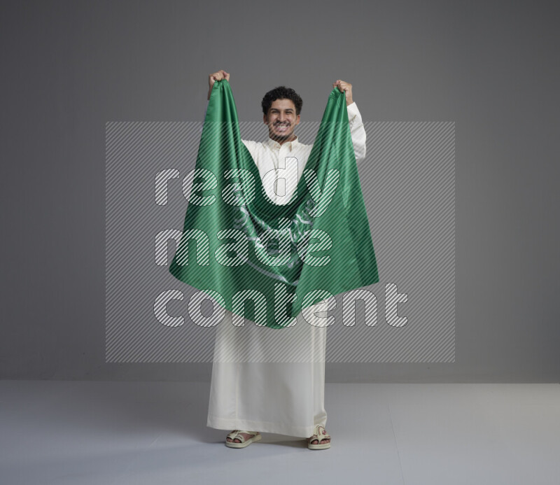 A Saudi man standing wearing thob holding big Saudi flag on gray background