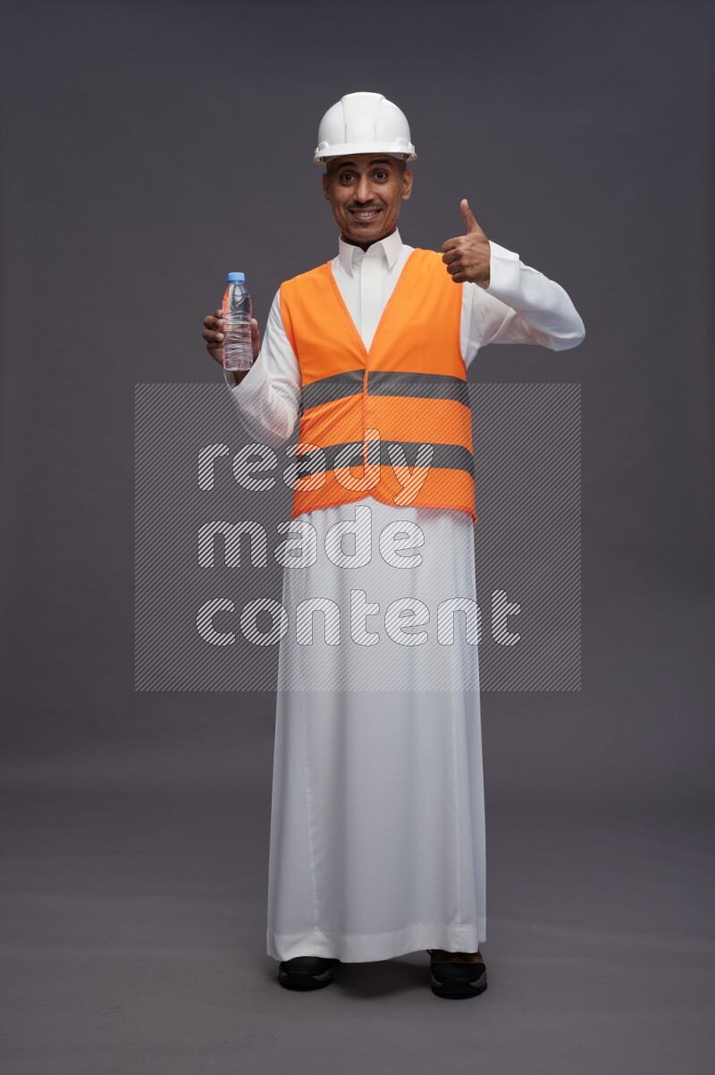 Saudi man wearing thob with engineer vest standing drinking water on gray background