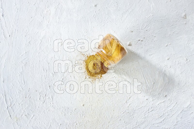 A flipped glass spice jar full of turmeric powder and powder fell out it on textured white flooring