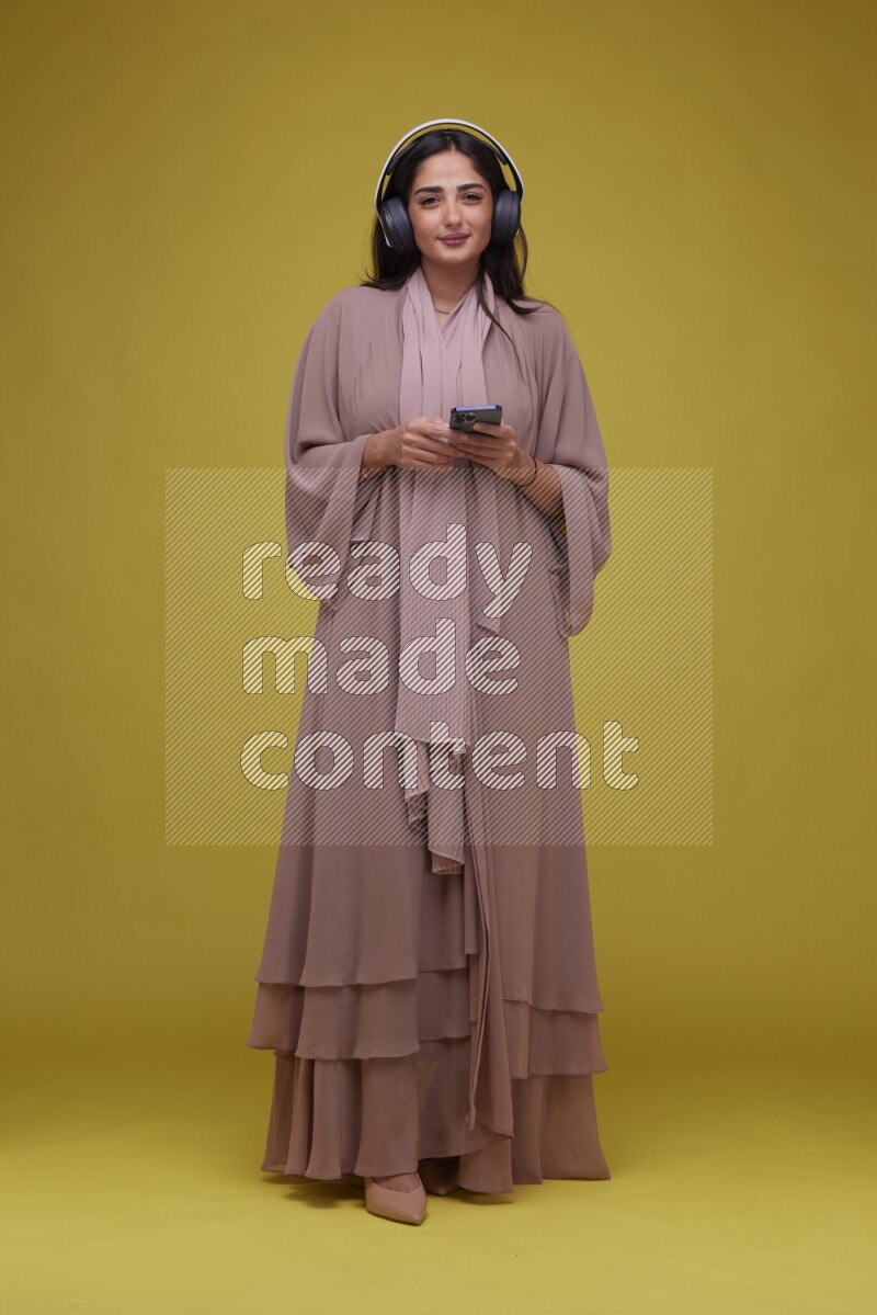 A Saudi woman Listing to Music on a Yellow Background wearing Brown Abaya