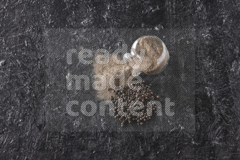 A flipped glass spice jar full of black pepper powder with pepper beads on textured black flooring