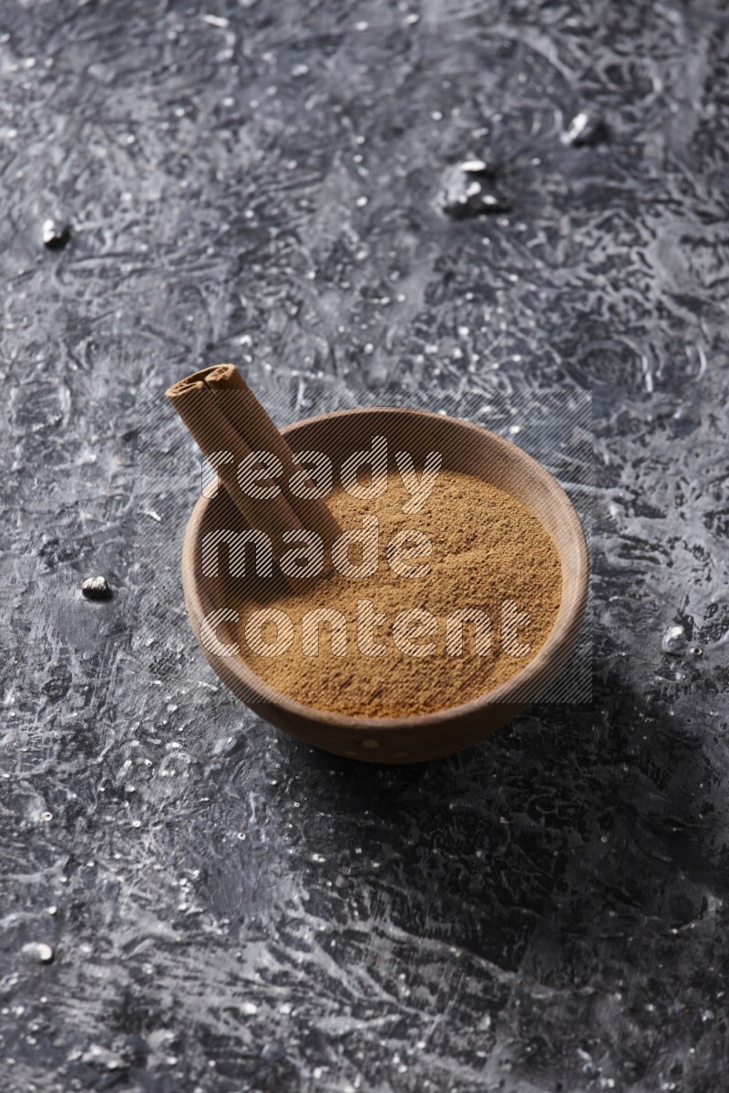 Wooden bowl full of cinnamon powder and a cinnamon stick on a textured black background