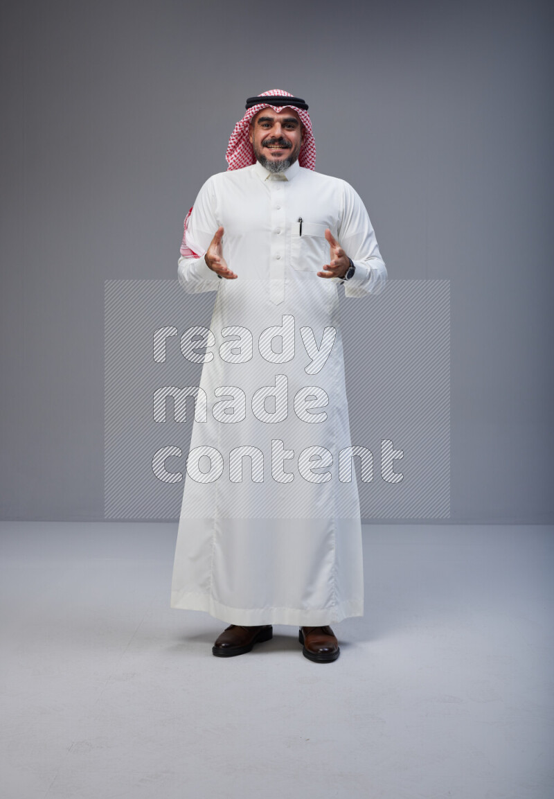 Saudi man Wearing Thob and red white Shomag standing interacting with the camera on Gray background