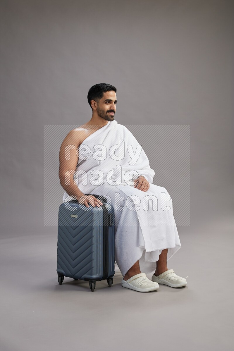A man wearing Ehram sitting on chair holding traveling bag on gray background