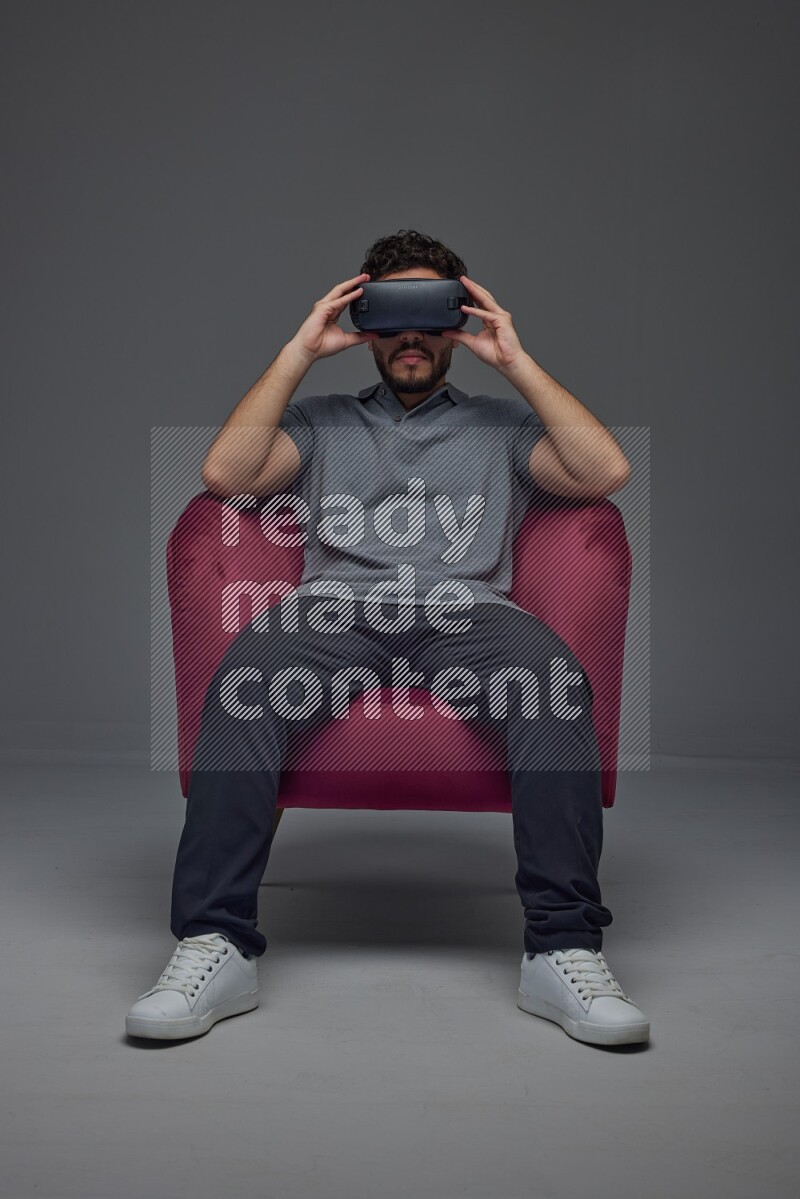 A man wearing casual and wearing VR while sitting on a burgundy chair eye level on a gray background