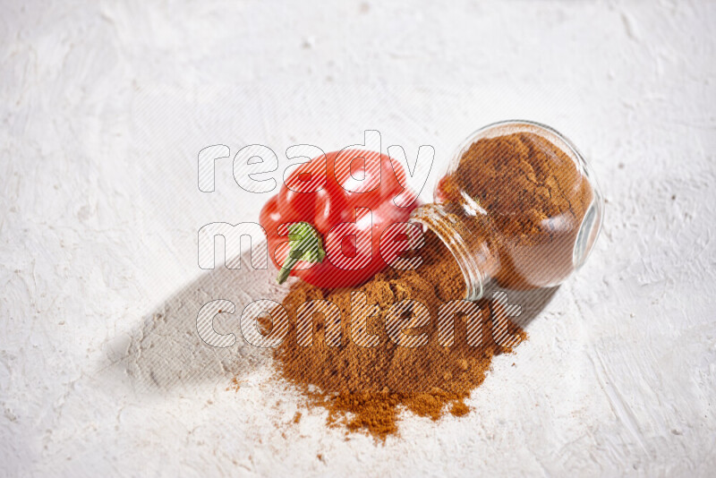 A glass jar full of ground paprika powder flipped with some spilling powder on white background