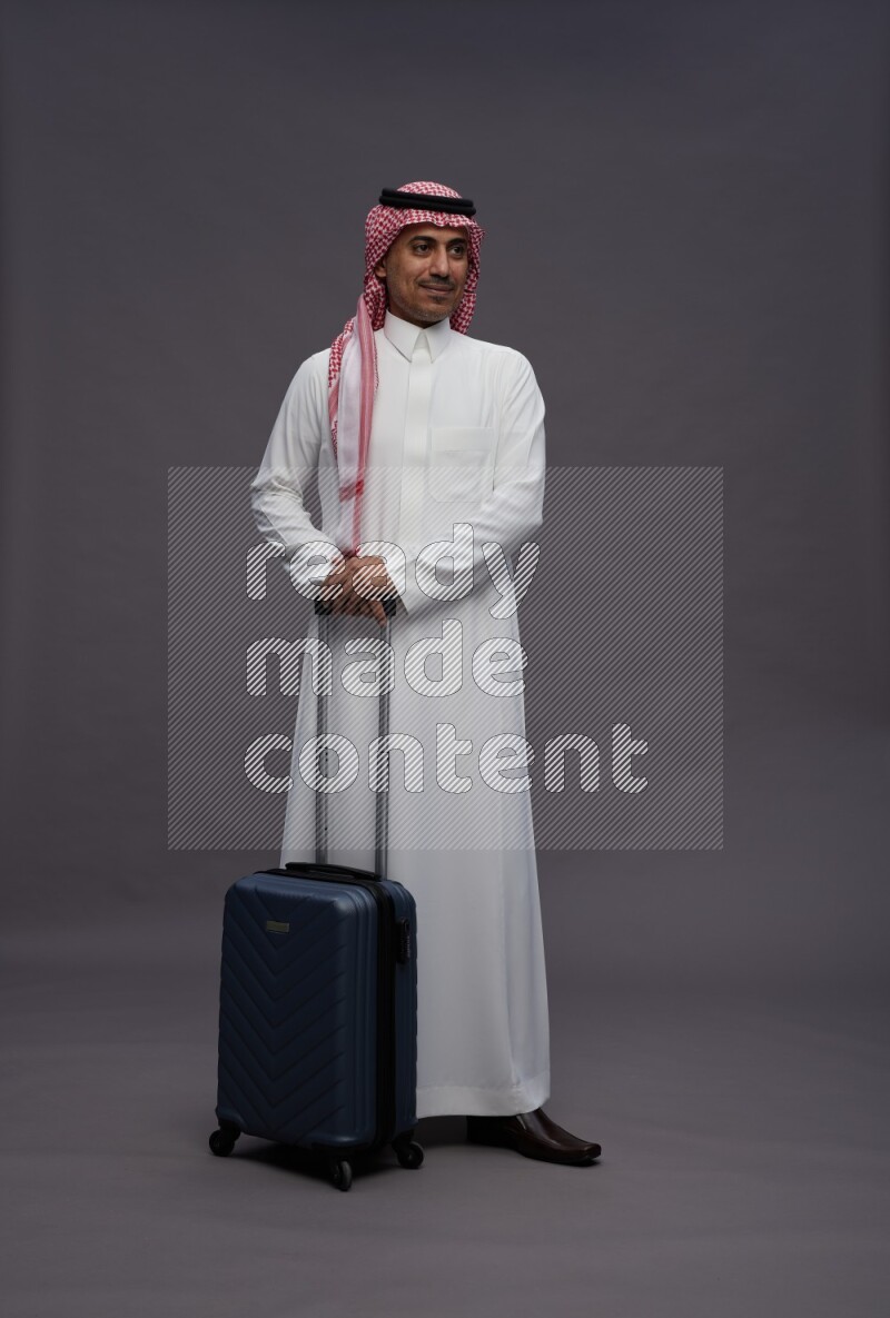 Saudi man wearing thob and shomag standing holding bag on gray background