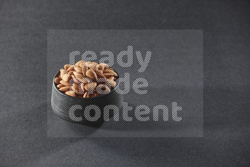 A black pottery bowl full of peeled almonds on a black background in different angles
