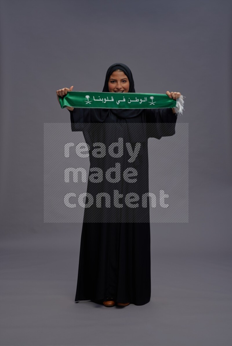 Saudi woman wearing Abaya standing holding Saudi flag on gray background