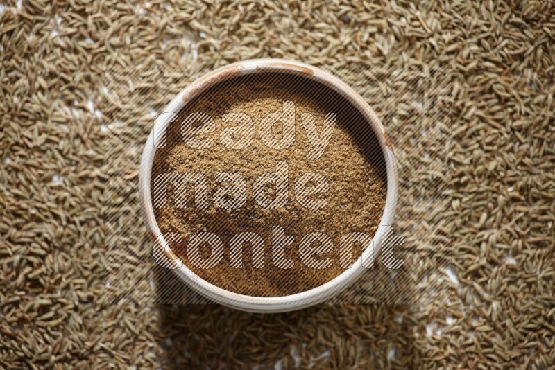 A beige bowl full of cumin powder with cumin seeds beneath it the bowl on a white flooring