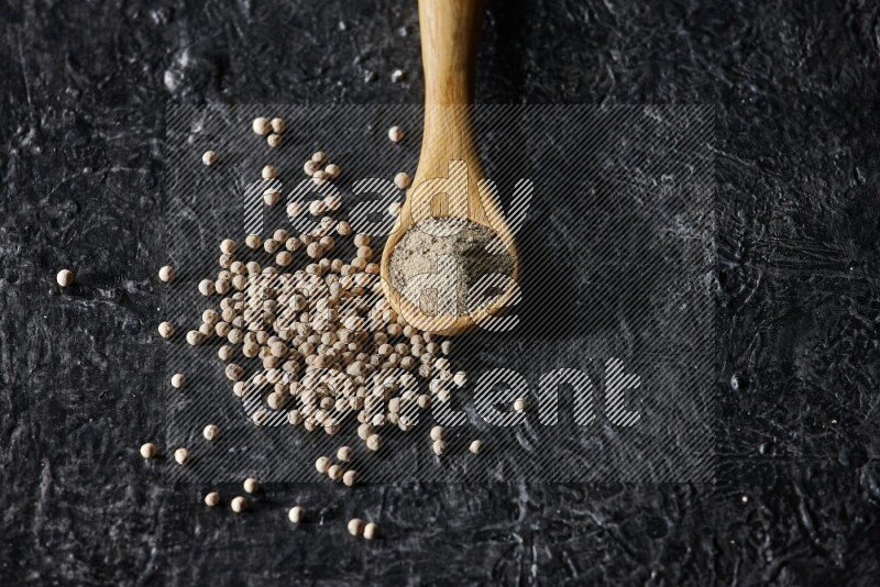 A wooden spoon full of white pepper powder with white pepper beads on textured black flooring