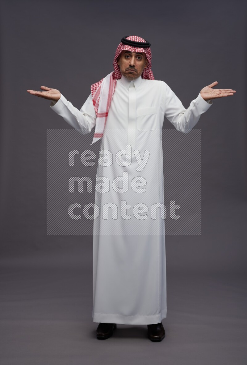 Saudi man wearing thob and shomag standing interacting with the camera on gray background
