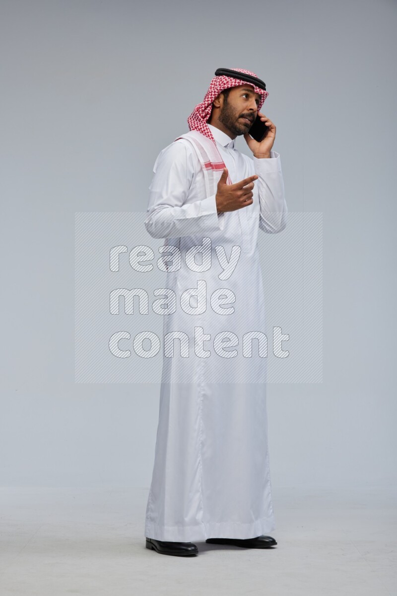 Saudi man Wearing Thob and shomag standing talking on phone on Gray background
