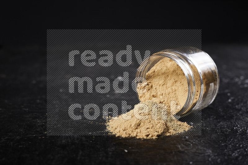 A glass jar full of ground ginger powder flipped with some spilling powder on black background