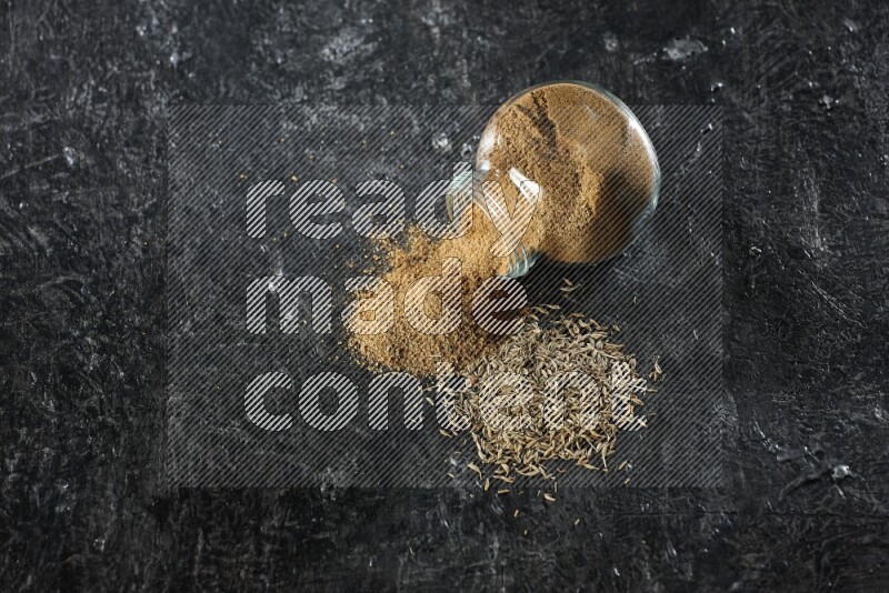 A flipped glass spice jar full of cumin powder with spilled powder and cumin seeds on a textured black flooring