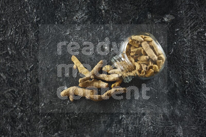 وعاء زجاجي للتوابل مقلوب وممتلئ بأصابع الكركم المجففة وبعض أصابع الكركم خارج الوعاء على خلفية سوداء