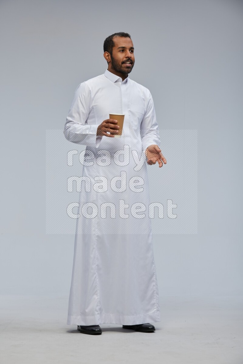 Saudi man wearing thob standing holding paper cup on gray background