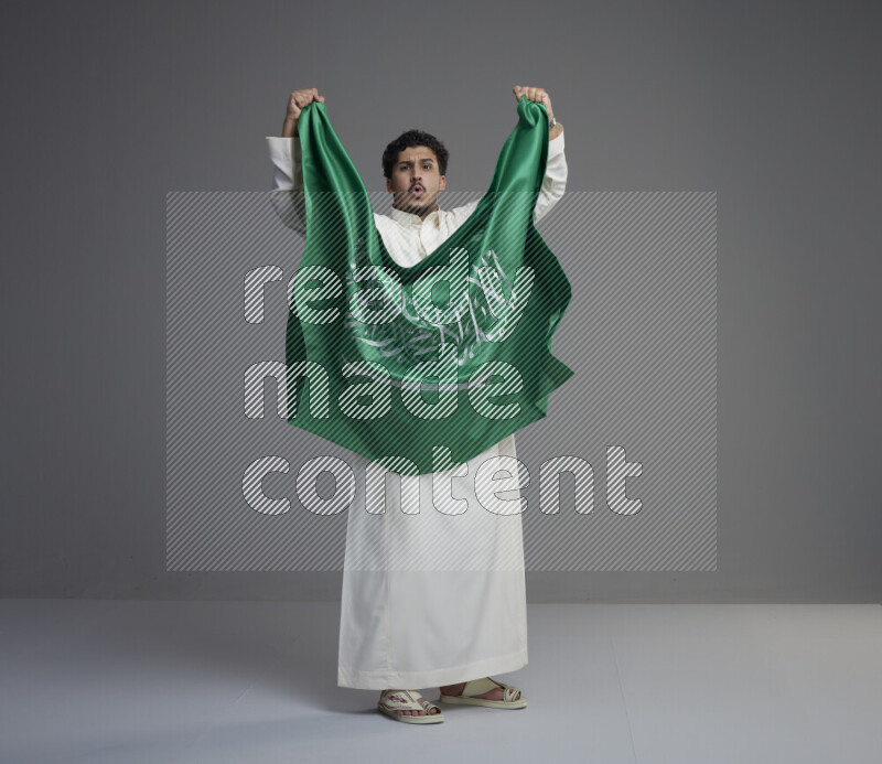 A Saudi man standing wearing thob holding big Saudi flag on gray background