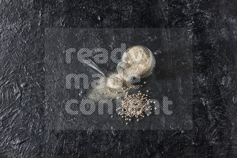 A flipped herbal glass jar and metal spoon full of white pepper powder with spilled powder and pepper beads on textured black flooring
