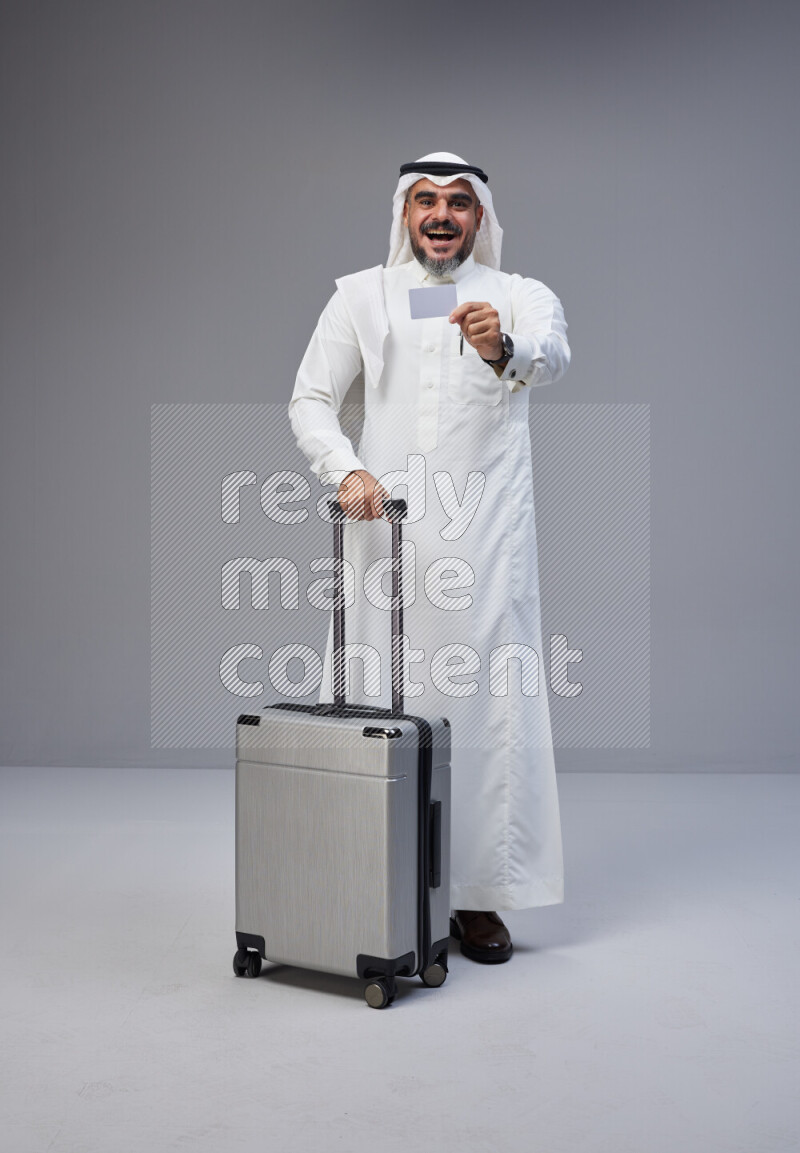 Saudi man wearing Thob and white Shomag standing holding Travel bag and ATM card on Gray background