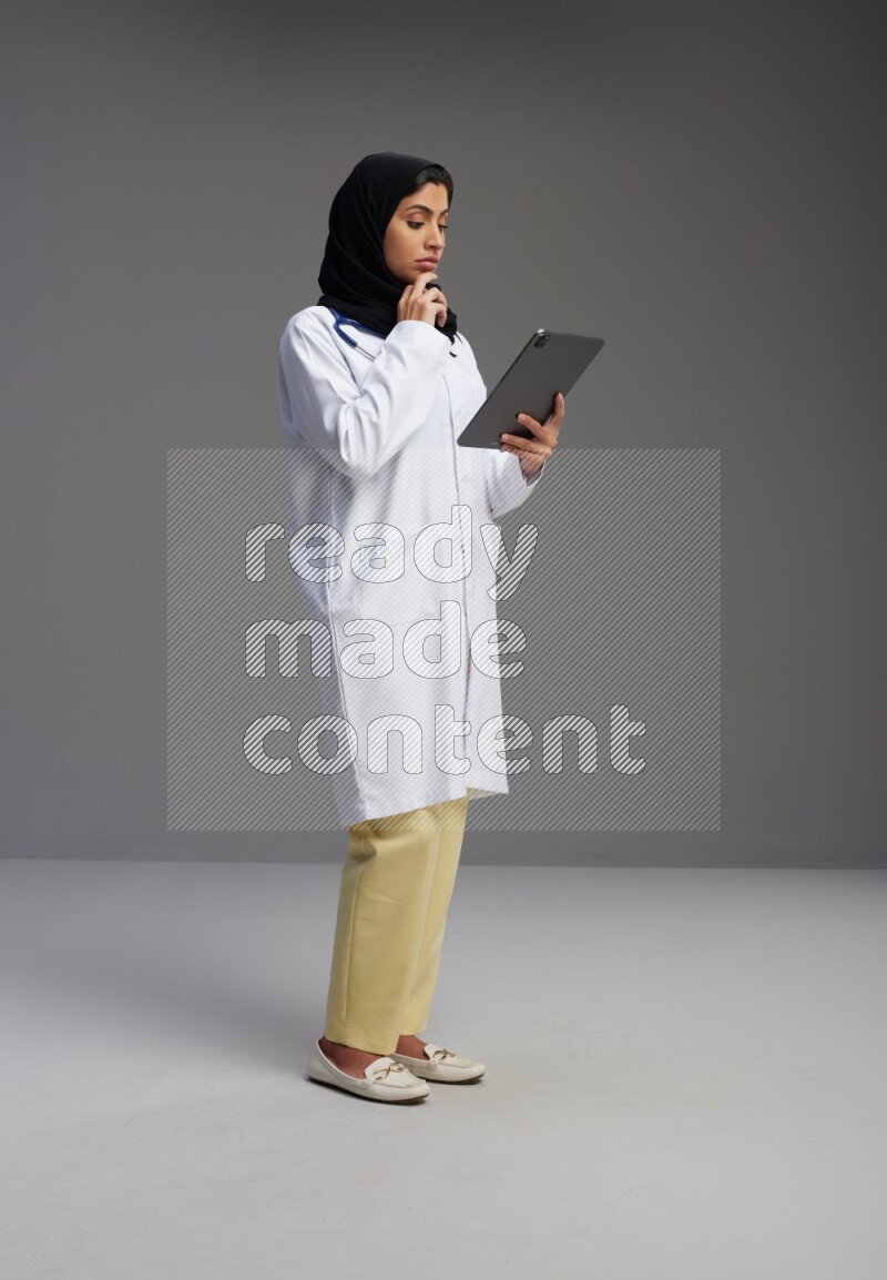Saudi woman wearing lab coat with stethoscope standing working on tablet on Gray background