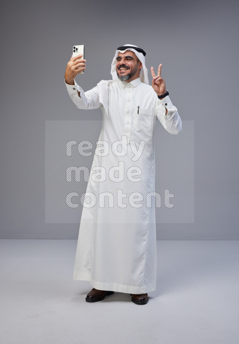 Saudi man Wearing Thob and white Shomag standing taking selfie on Gray background