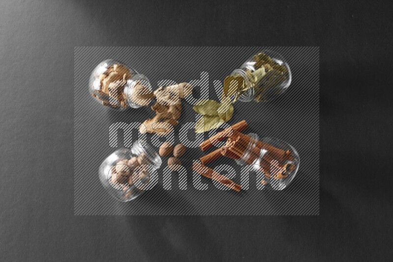 4 glass spice jars full of laurel bay leaves, ginger, cinnamon and nutmeg on black flooring