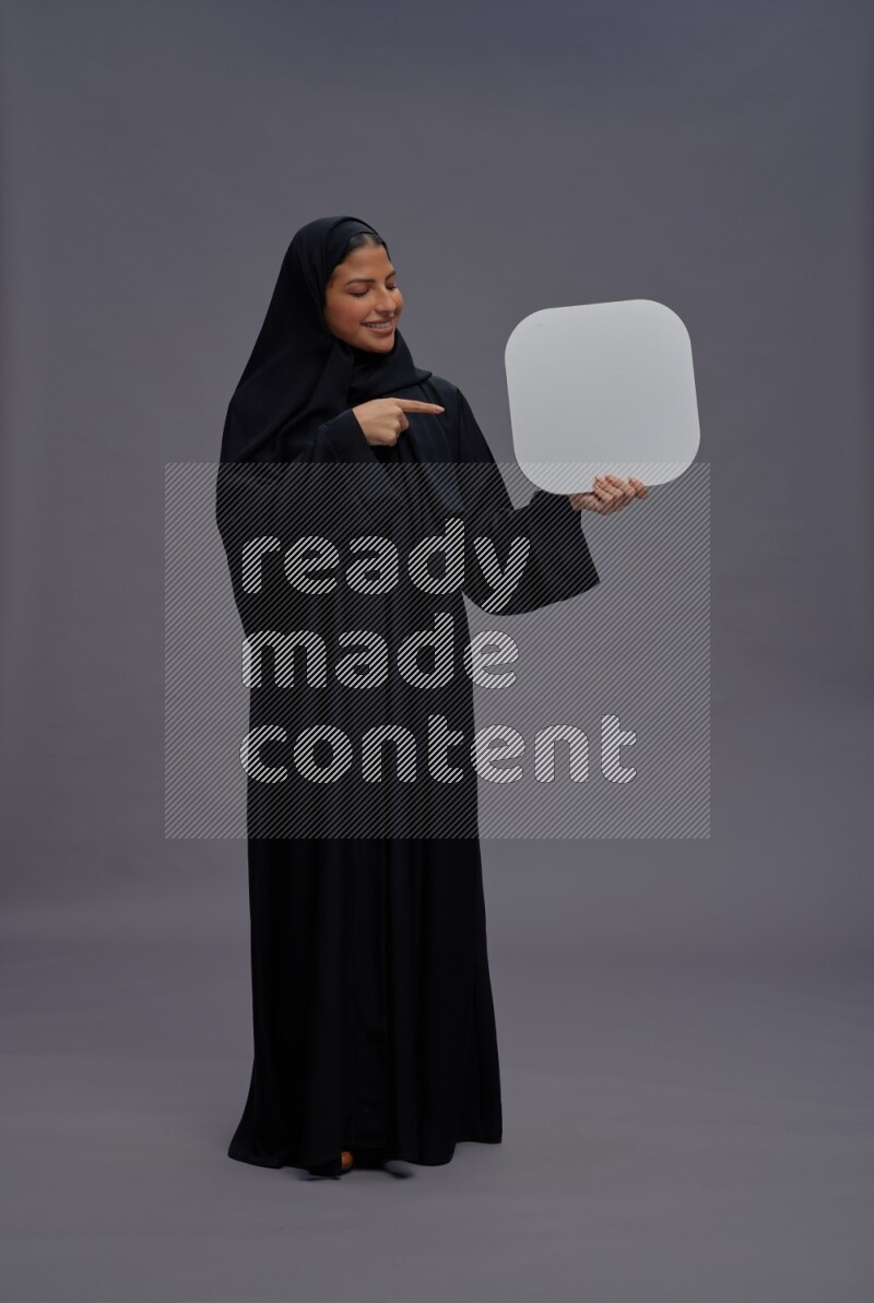 Saudi woman wearing Abaya standing holding social media sign on gray background